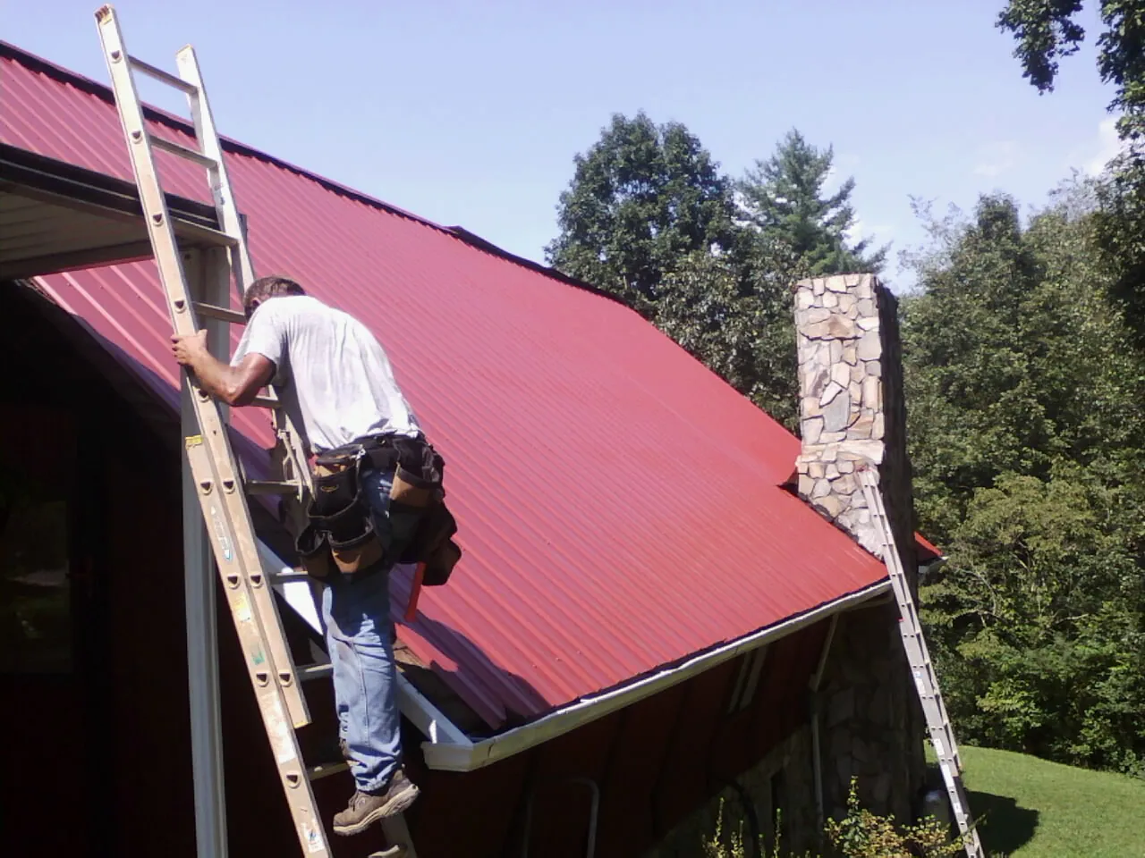 Roofer on a ladder beside a red metal roof and stone chimney — Soffit & Fascia Repair by Roofing Brothers in Harper